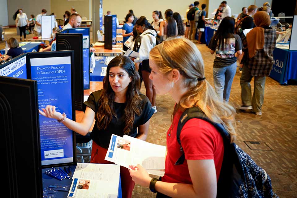 A student speaks with a presenter at a booth during an academic fair. There is a poster that reads "Didactic Program in Dietetics"