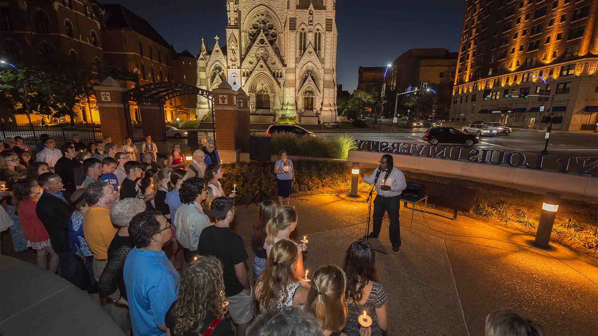 Dr. Jonathan Smith stands in front of a group of people at night, at the corner or Lindell and Grand, with the Saint Louis University sign in the background.