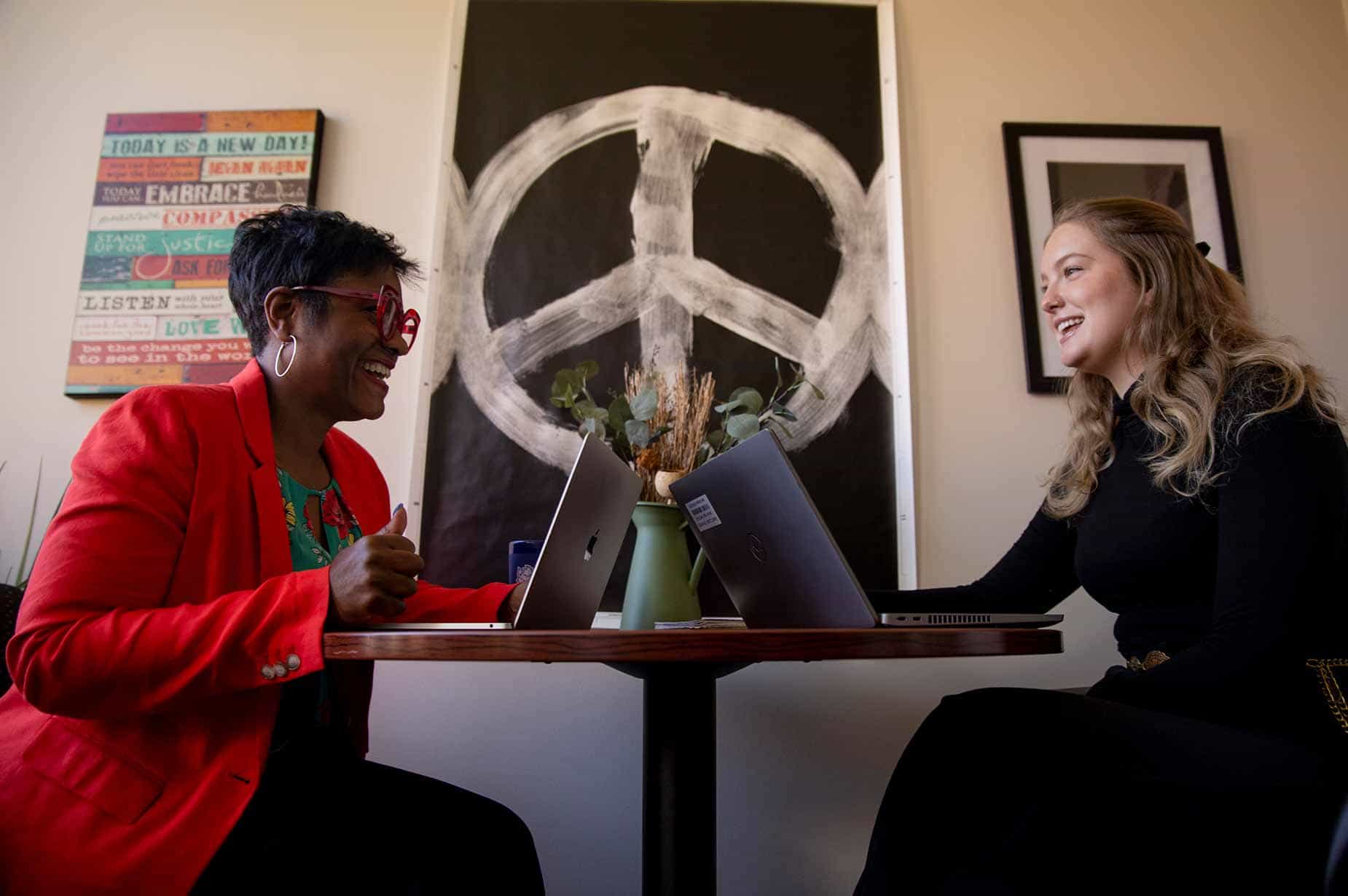 A female professor works with a group of students while sitting at a table. They are looking over paperwork and chatting.