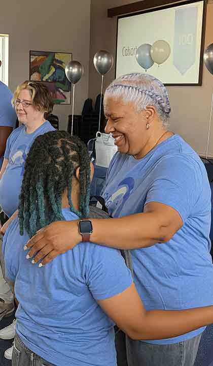 A woman hugs a younger woman during a celebration. Both are wearing matching STL Teaching Fellows shirts.