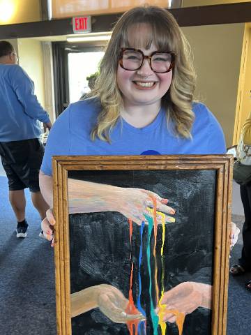 A smiling woman holds a framed painting, which depicts a hand pouring streams of multiple colors of paint onto two hands below.