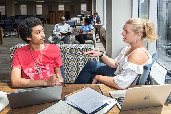 Two students, a male and a female, are speaking at a table that overlooks the courthouse in the Vincent C. Immel Law Library.