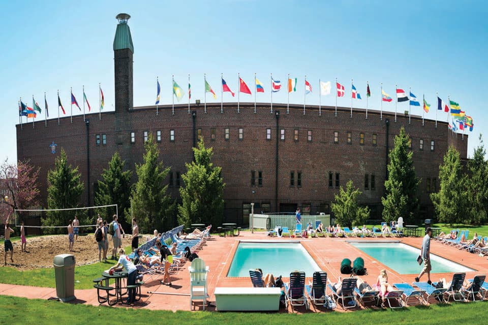 People play sand volleyball and lie on chairs next to two swimming pools.