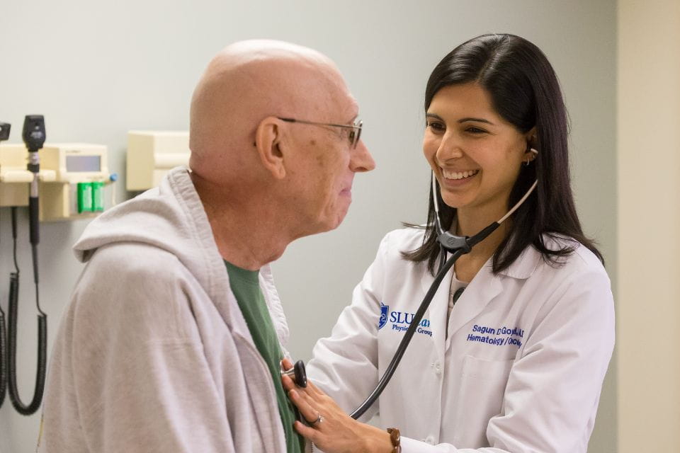 A physican smiles at her patient while listening to his heart with a stethoscope inside a hospital exam room.