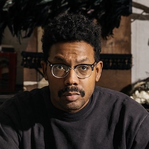 A shoulder-length photograph of a man with textured black hair, a moustache, and wearing black-rimmed glasses. His face is dramatically illuminated from the left and he looks intently straight ahead, his right eyebrow slightly raised. He wears a black crew-neck shirt. The room in the background is out of focus.