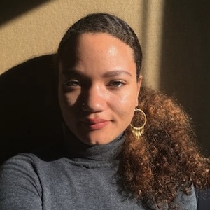 A shoulder-length photograph of a young adult woman with auburn-tinted black hair pulled back in a pony tail that drapes across her left shoulder. She wears a heather-gray turtleneck sweater and large gold hoop earrings. She stands against a caramel-colored background. Golden light dramatically illuminates the left side of her face and leaves the right side in shadow.