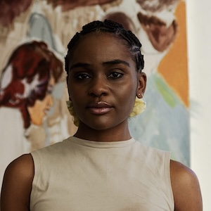 A shoulder-length photograph of a young adult woman with plaited black hair drawn into a light yellow band behind her head. She wears a sleeveless cream-colored top and stands looking slightly down at the camera with an intent gaze. In the background, out of focus, is a painting with a woman in profile who is positioned just to the right of the subject's head. 