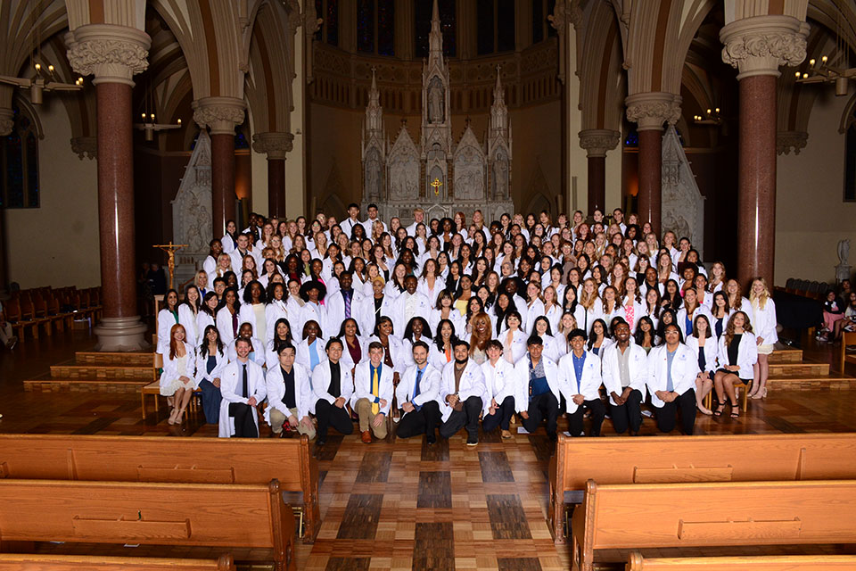 A group of students wearing white coats pose for a photo inside a dimly lit church.