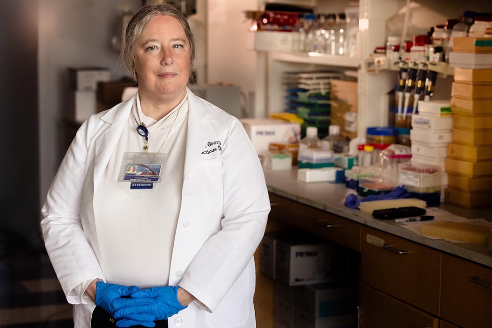 A woman wearing a white lab coat and blue gloves poses for a photo in a dimly lit lab. Vials of tubes and boxes are on shelves and a counter in the background.