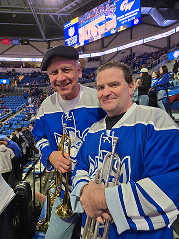 Billiken Pep Band trumpeters Anselman and Huether An man in a hat and a brown-haired man wear blue-and-white hockey jerseys and hold trumpets.