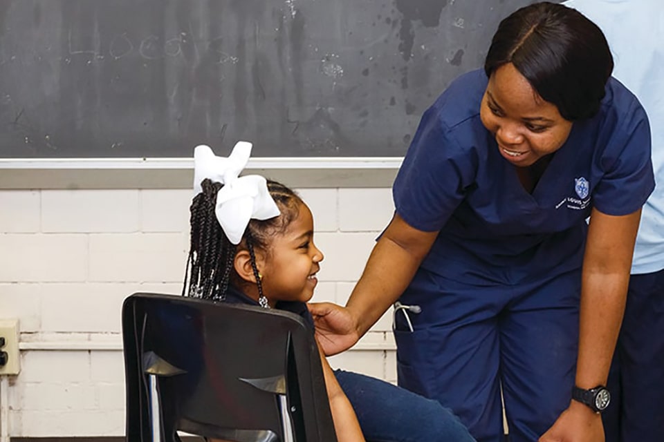 A student wearing nursing scrubs smiles while talking to a young girl in a classroom.