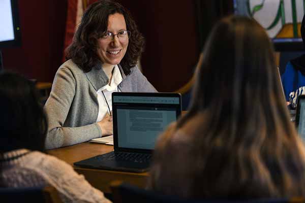 A female faculty member speaks with students while sitting around a table. The students have laptops open to work on.