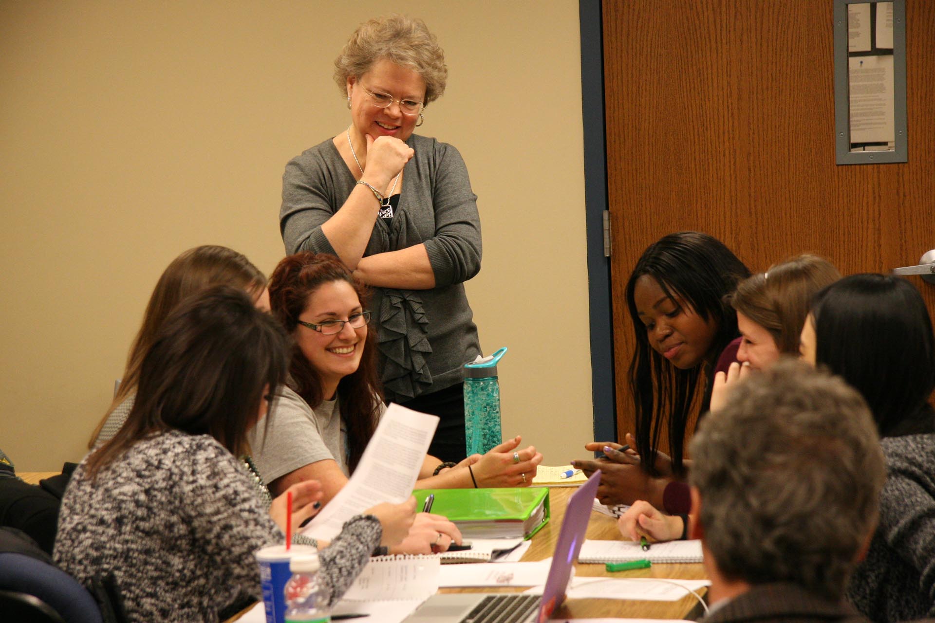 Students sit at a table with laptops, binders and water bottles during a discussion while a professor looks on. 