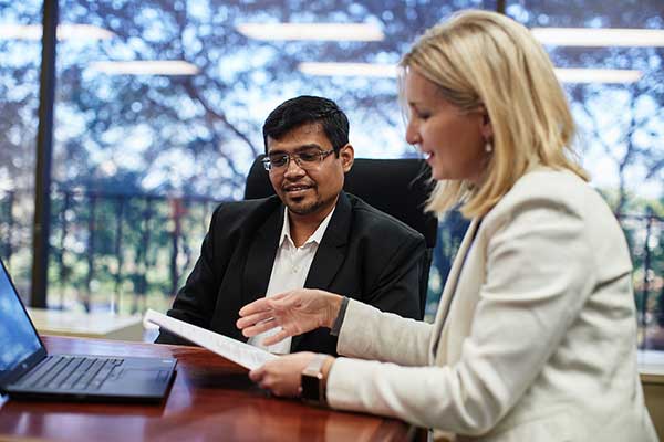 A staff member in a white blazer holds a piece of paper and goes over something with a student in glasses with a black suit coat on at a conference table.