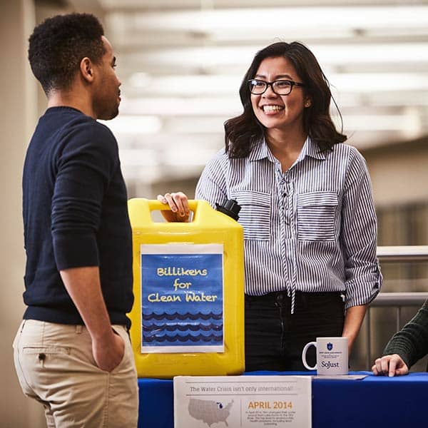 A female student smiles while speaking with another male student and attending a table in Busch Student Center. A large yellow plastic jug sits on the table with a label that reads “Billikens for Clean Water.”