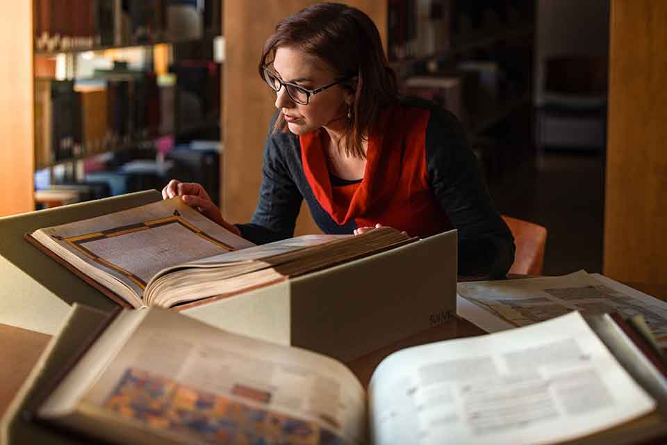 A woman leans over three illuminated manuscripts that are spread across a table top.