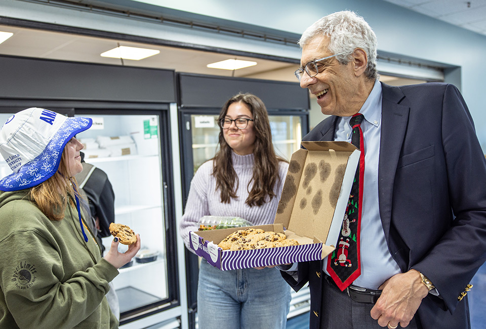 Pestello holds a box of cookies and offers one to a female student wearing a SLU bucket hat while a woman in glasses looks on.