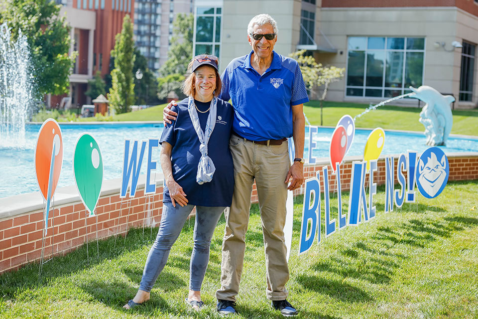 Fran and Fred Pestello wear casual clothes and stand by the Dolphin Pond on SLU's campus. Colorful signs line the pond.